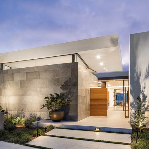 A modern, minimalist house exterior with large stone walls, wooden elements, sleek lighting, and a landscaped front yard at dusk.