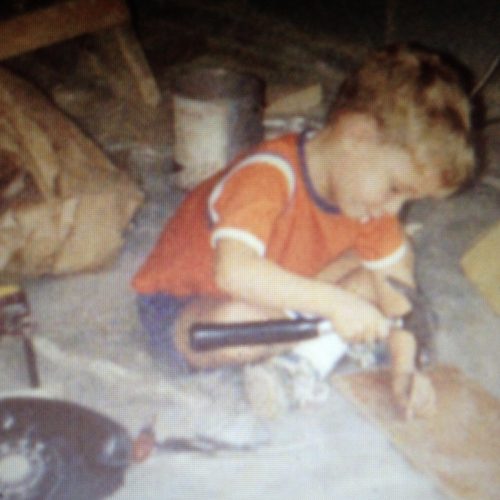 This image shows a young child in an orange shirt kneeling on the floor, focused on hammering a nail into a piece of wood, surrounded by various tools and materials.