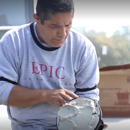 A man wearing an "EPIC Ceramic Stone" sweatshirt applies mortar to a trowel, focusing intently on his work.